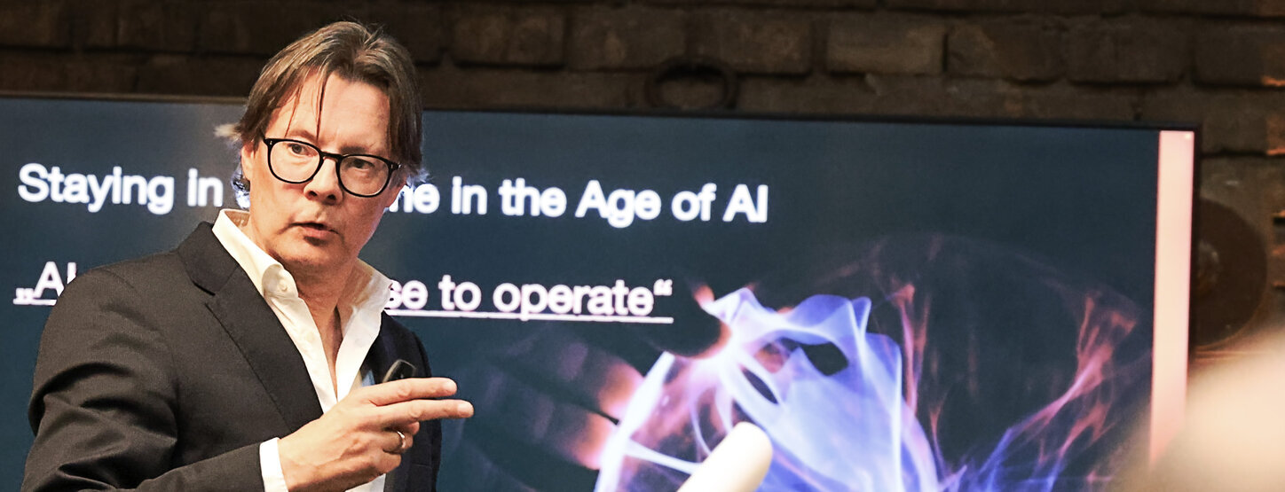 Markus Webhofer im Vordergrund, im Hintergrund ist ein Bildschirm zu sehen; Startseite mit Thema Staying in the game in the Age of AI