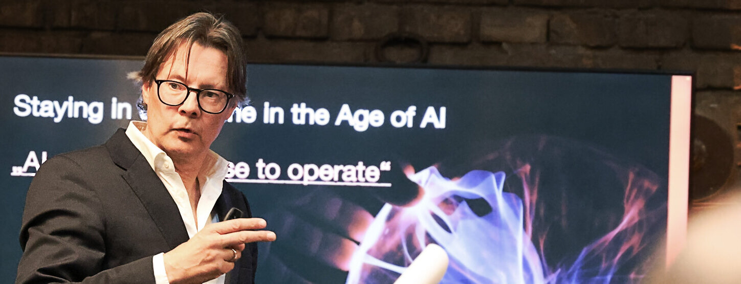 Markus Webhofer im Vordergrund, im Hintergrund ist ein Bildschirm zu sehen; Startseite mit Thema Staying in the game in the Age of AI